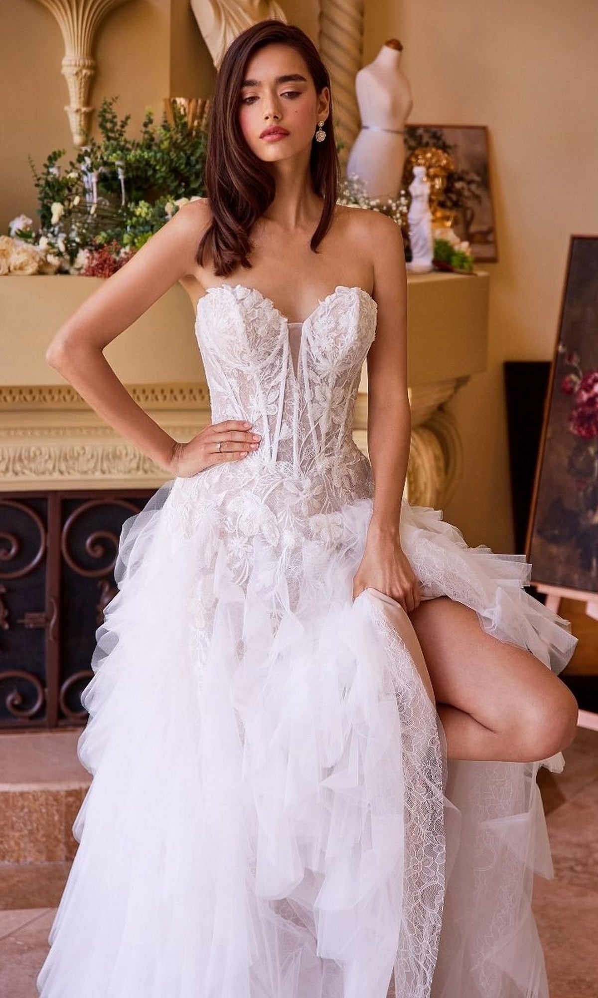 Wearing the Andrea and Leo WL016, a woman poses indoors in a strapless wedding gown with a lace-applique corset bodice, one hand on her hip and the other lifting the ruffled tulle A-line skirt to reveal her leg.